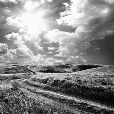 Paysage en noir et blanc avec un chemin sinueux sous un ciel nuageux lumineux.
